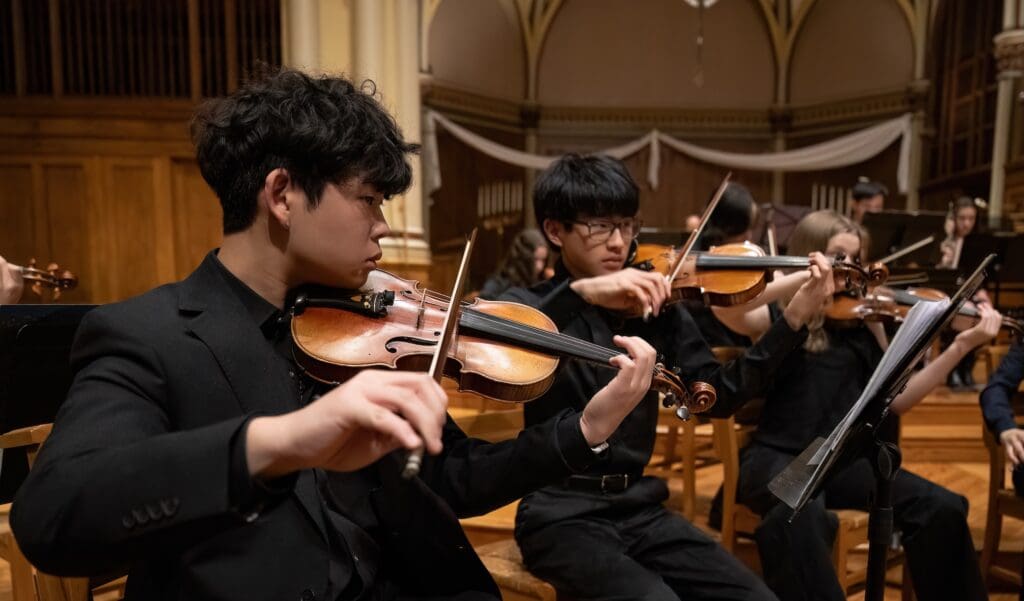 A group of young musicians in formal attire play violins during an orchestral performance in a concert hall.