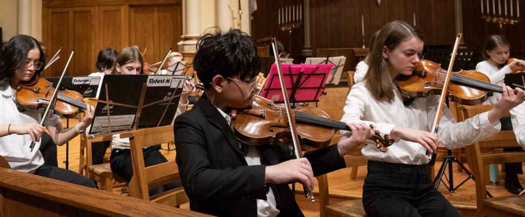 A group of young musicians in formal attire play violins and read sheet music during an indoor orchestra performance.