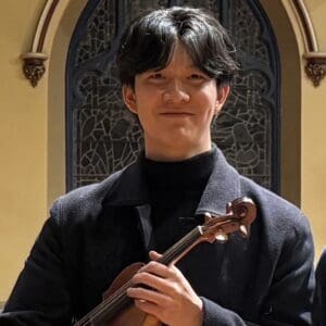 Young People's Philharmonic Person holding a violin, standing indoors in front of a decorative arched window, wearing a dark coat and smiling slightly.