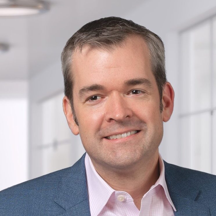 A man in a blue suit jacket and light pink shirt stands smiling in a modern office hallway with glass walls and overhead lights.