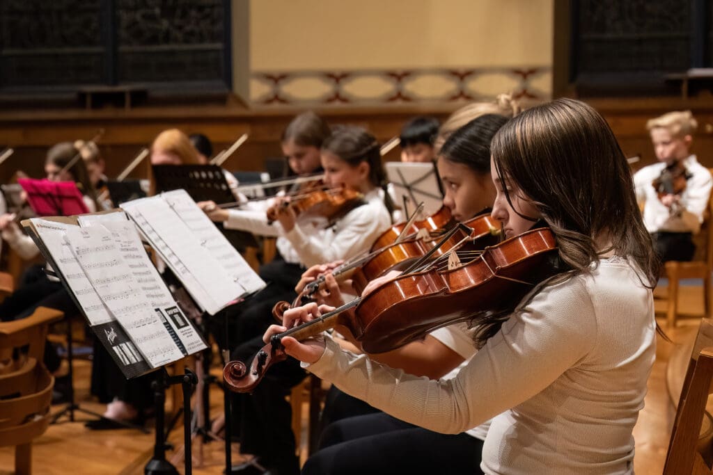 A group of young musicians in white shirts play violins in an orchestra, reading sheet music during a performance in a concert hall.
