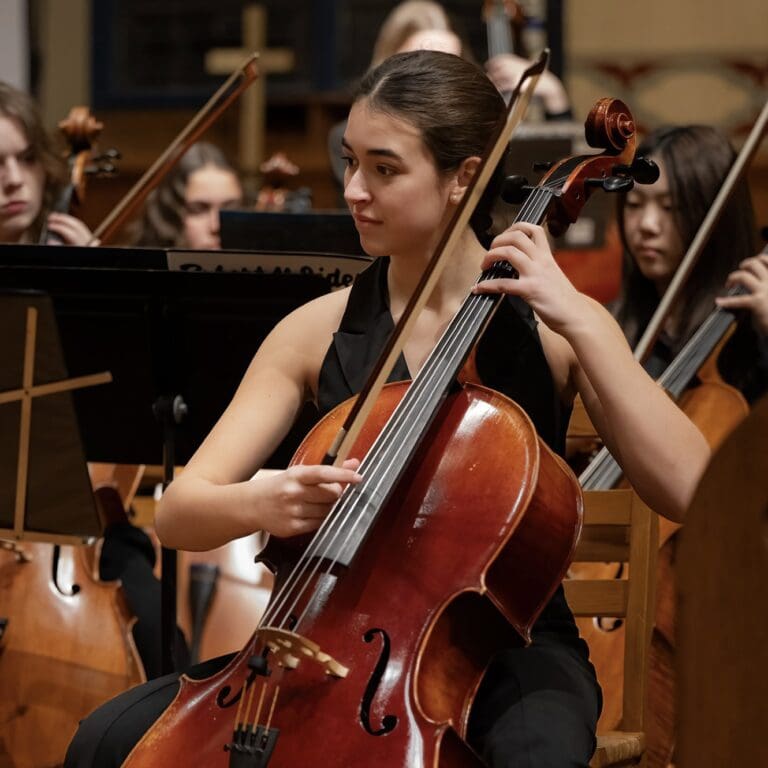 A young woman plays the cello in an orchestra, focused on her performance, surrounded by other musicians and musical instruments.