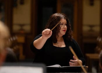 A conductor in black attire leads an orchestra during a performance, holding a baton with musicians and music stands visible in the foreground.