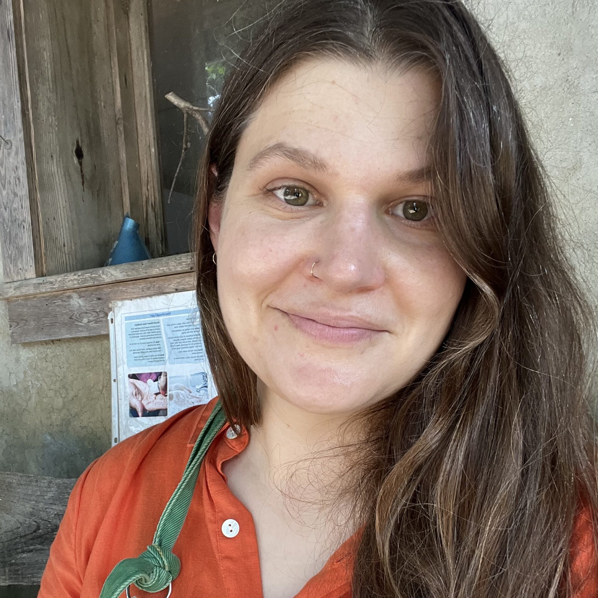 A person with long brown hair and a nose ring, wearing an orange top and green apron, stands in front of a rustic wooden window and wall.