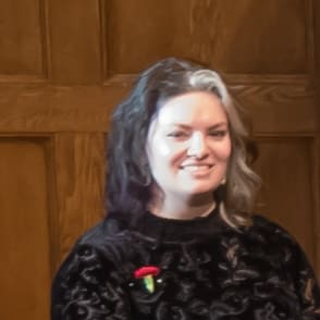 A person with shoulder-length hair, partly dark and partly light, wearing a black top with a red and green pin, stands in front of a wooden paneled background.
