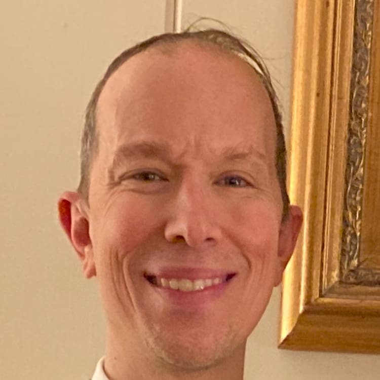 A man with short hair smiles at the camera, standing indoors near a gold-framed picture.