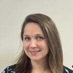 A woman with long brown hair and a slight smile poses in front of a plain, light-colored wall.