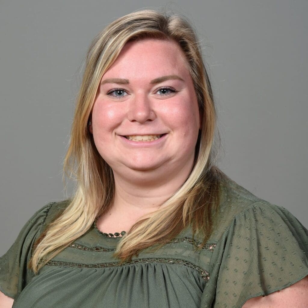 A woman with long blonde hair, wearing a green blouse, smiles at the camera in front of a plain gray background.
